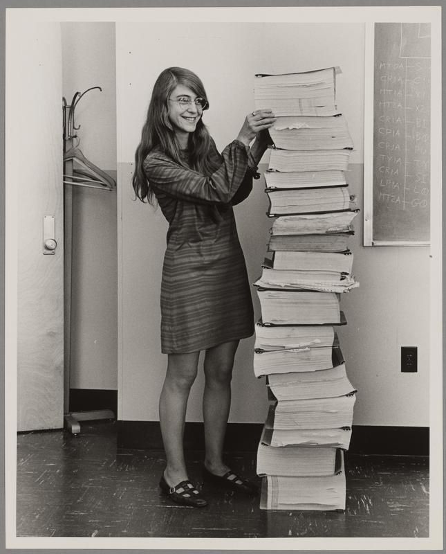 Hamilton has long hair, glasses and wearing a dress. A stack of papers almost as tall as she is are next to her